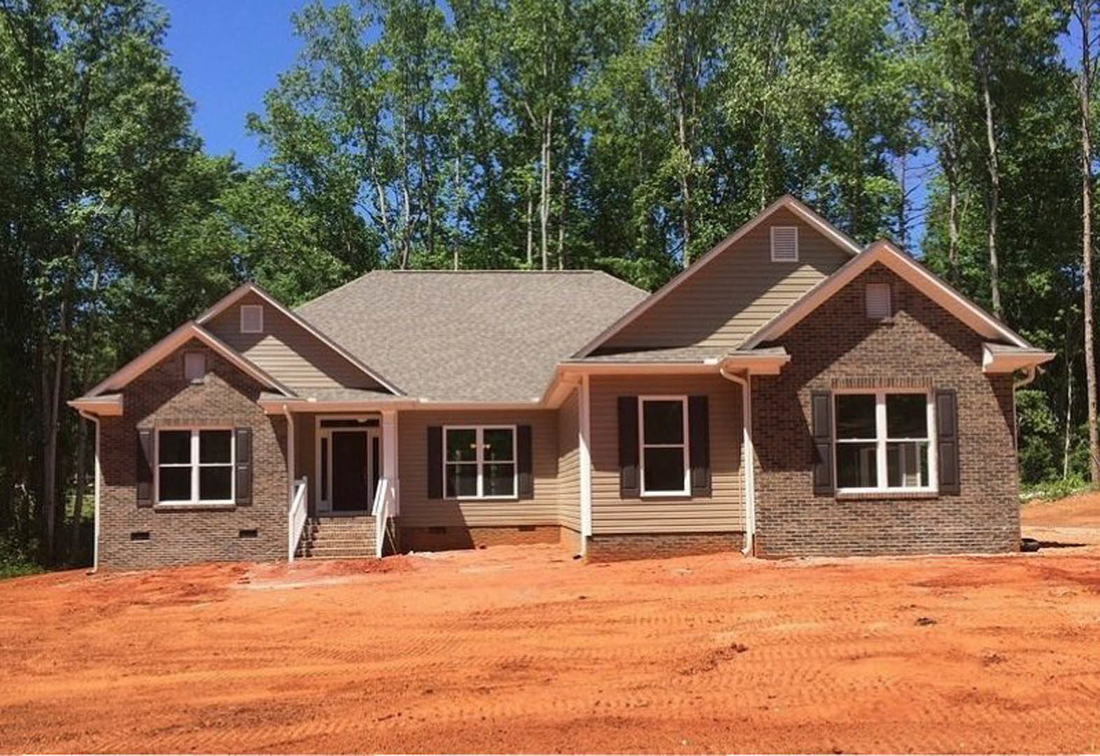 Framed house under construction with white window frames, unfinished roof, dirt yard with tire tracks, surrounded by mature trees