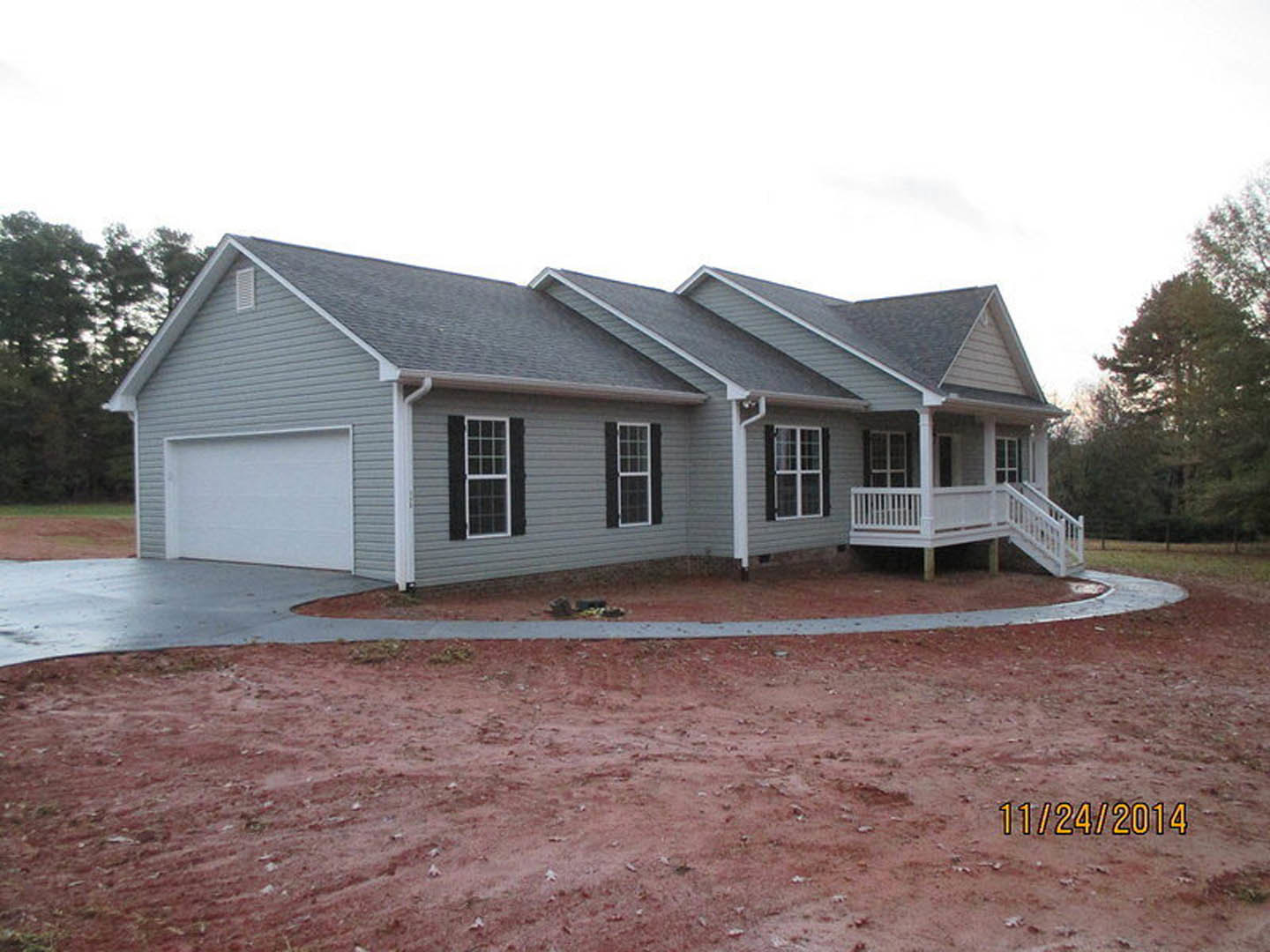 Grey siding with white trim, white porch railing, white-framed windows, attached white garage door, dirt yard, leafy trees, blue sky