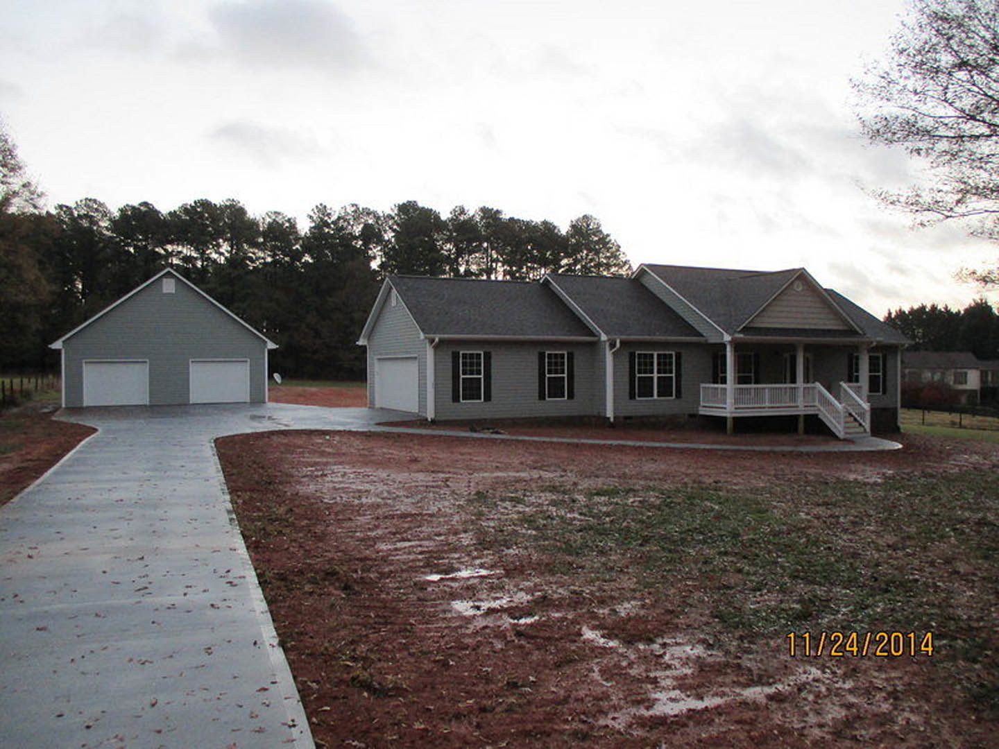 Grey two-story house with attached garages, white porch railing, large windows, and a paved driveway bordered by a dirt field with a puddle.