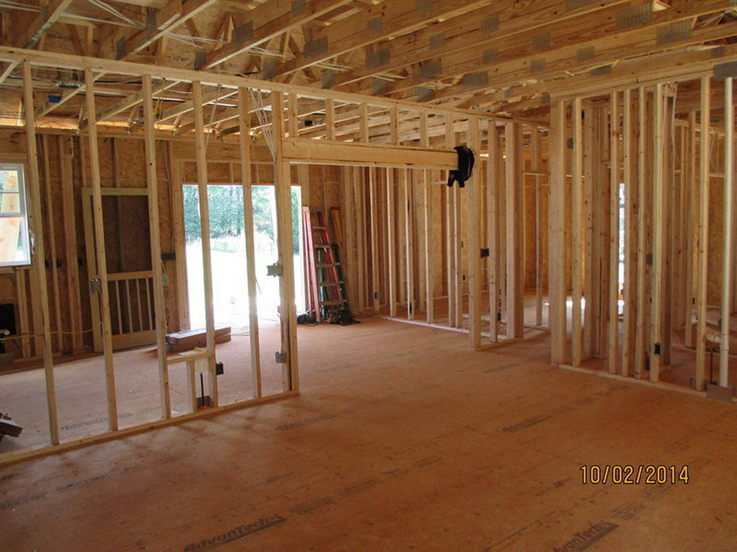 Wood-framed room under construction with exposed beams, open doorway, ladder, and unfinished flooring