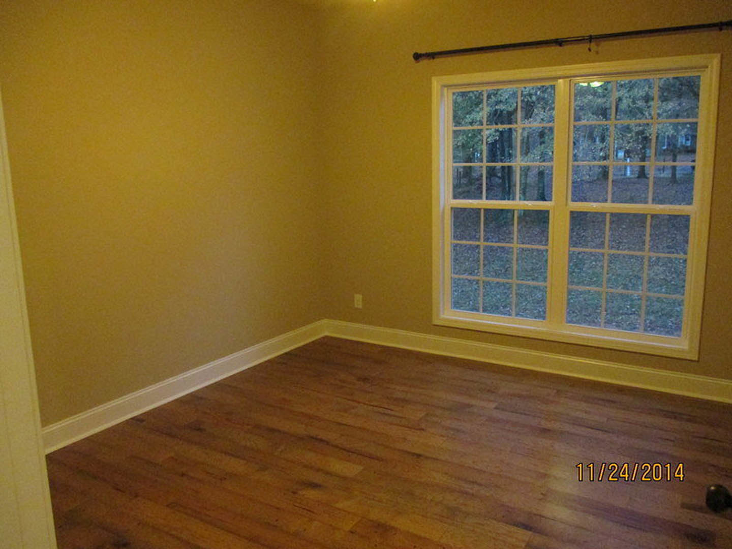 Sunlit room with hardwood flooring, large window framed by a curtain rod, view of leafy trees outside, neutral walls, and a small black decorative object on the floor.