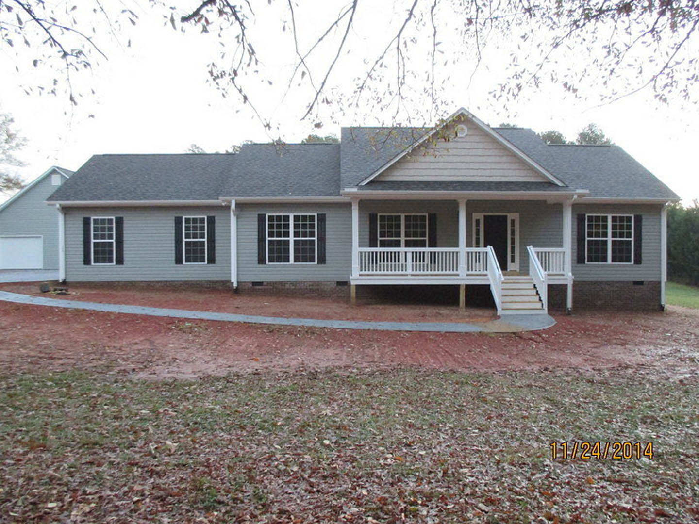 Gray siding house with covered porch, white railing, large windows, and leafy tree branches in foreground over green lawn.