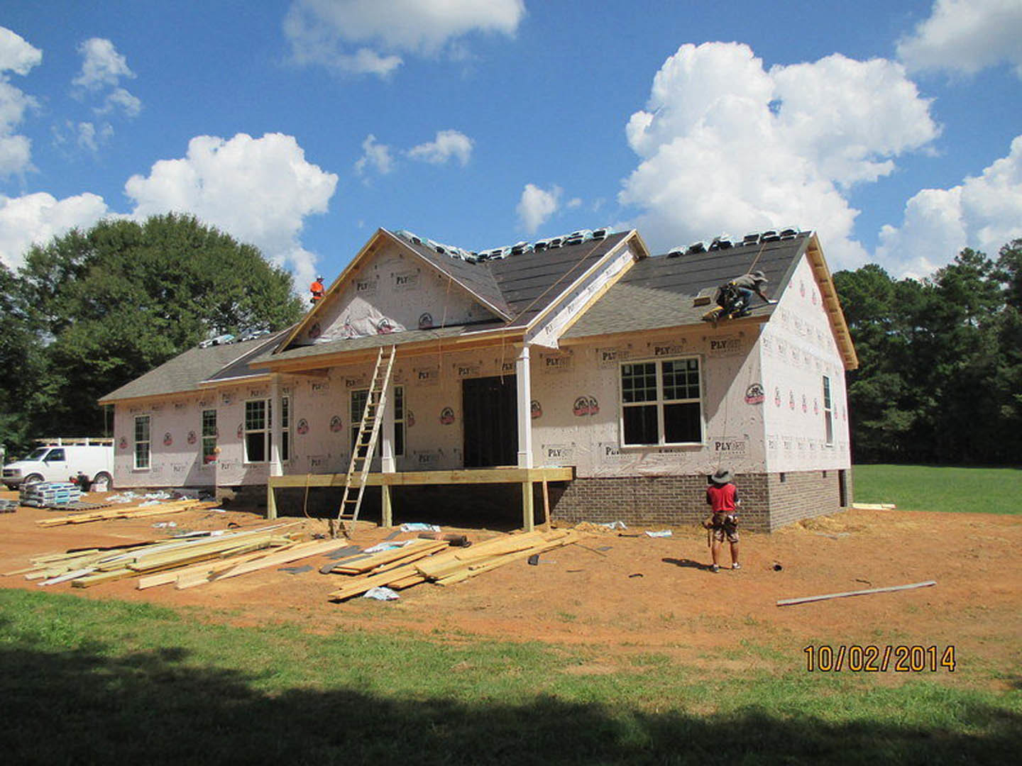 Wood-framed house under construction with exposed beams, ladder leaning against exterior, white van parked nearby, man in red shirt standing on site, grassy lot and trees in