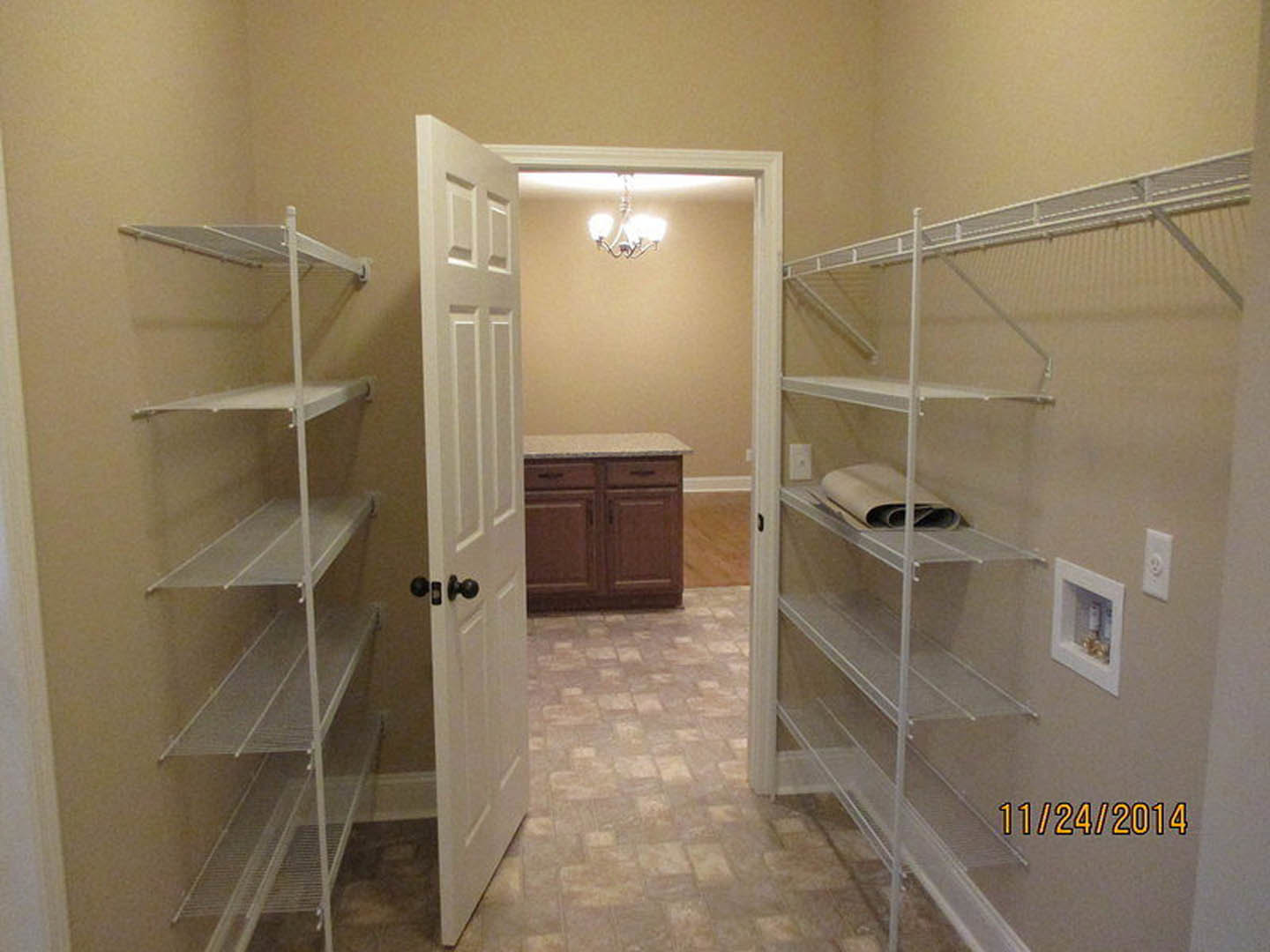 Open door leading to a room with white plaster walls, glass shelving displaying gold objects, and cabinetry; kitchen counter and rolled carpet visible.