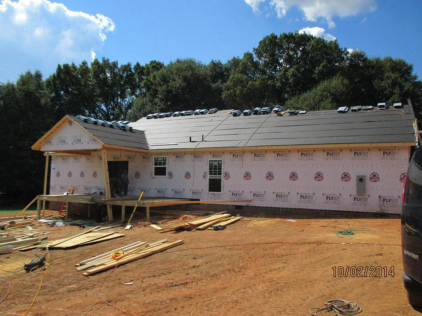 Framed wooden house under construction with exposed lumber, partially finished roof, white window frame, cars parked nearby, cloudy sky, and trees in background