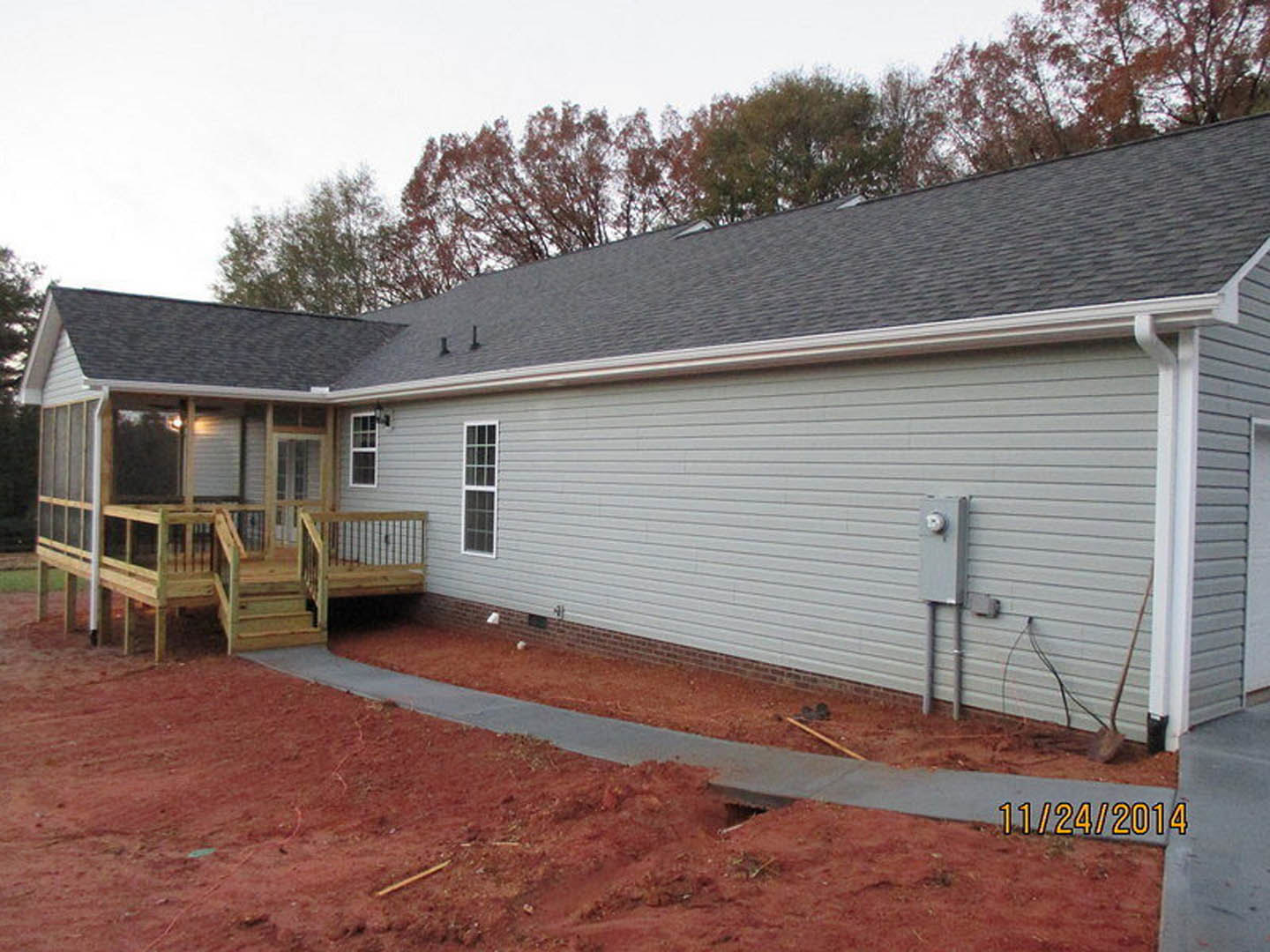 Two-story home with light siding, wooden deck and porch featuring railings, walkway leading to entrance, large windows, shingled roof, mature trees in background.
