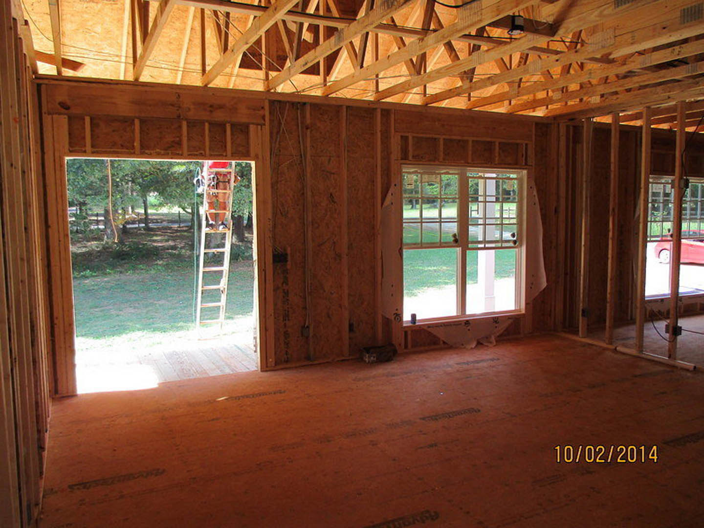 Sunlit room with exposed wooden ceiling beams, wood plank flooring, large window overlooking grassy yard, and a cat perched on a ladder.