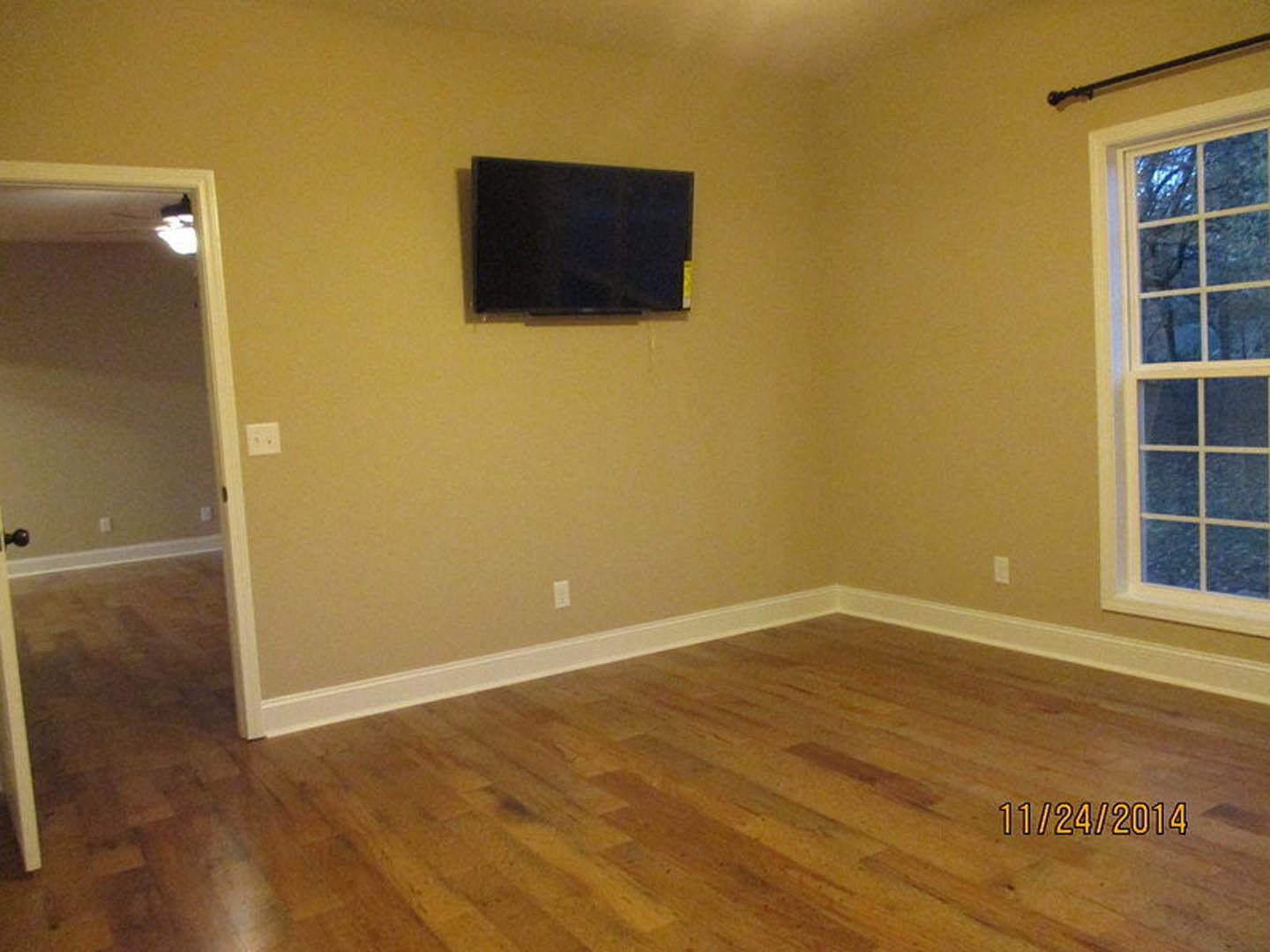 Hardwood floor living space with flat screen TV mounted on white wall, open door, white-framed window, and wall outlet with two switches