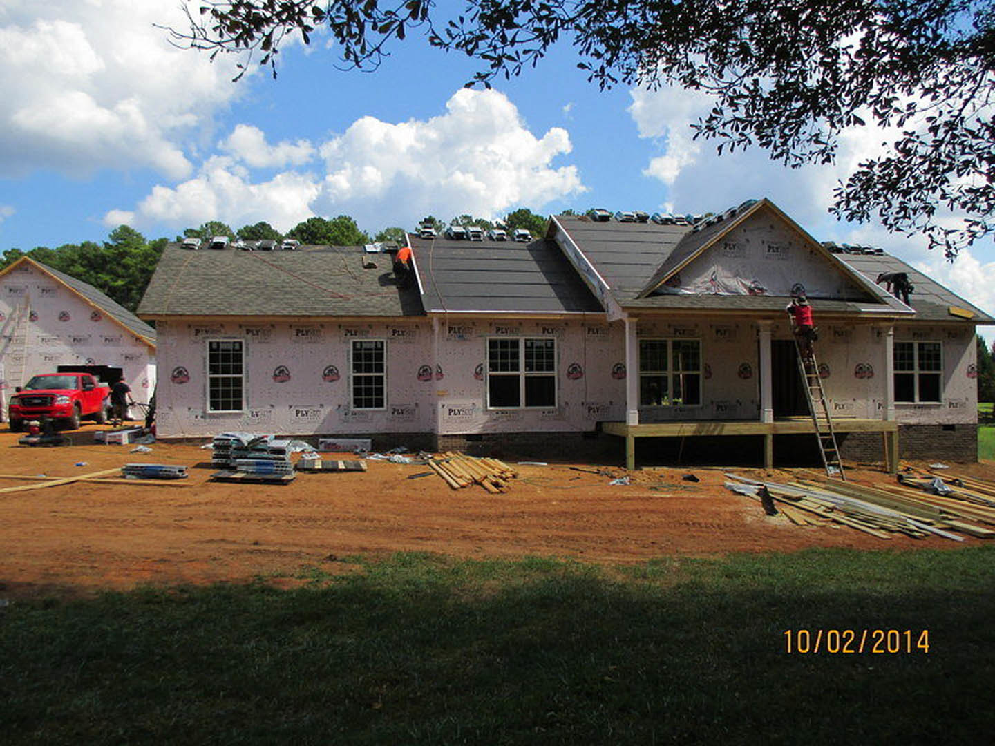 Partially constructed house with exposed roof panels, pile of lumber on dirt, red truck parked nearby, white pole and wooden table in foreground, large window installed, trees and