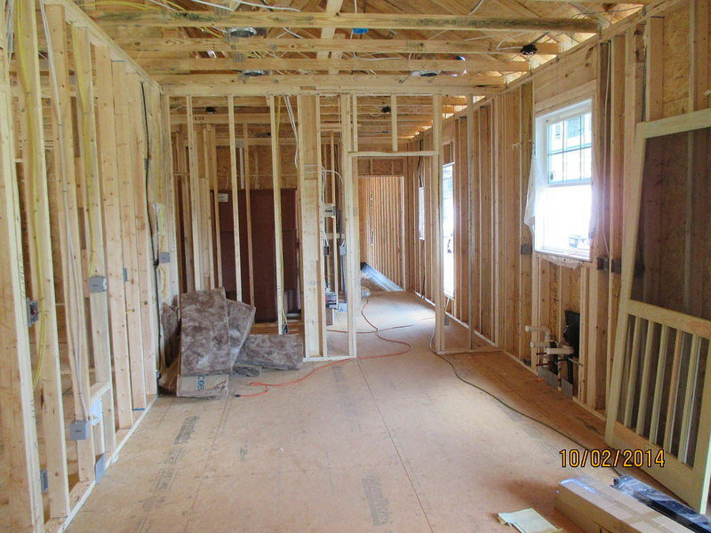 Wood-framed room under construction with exposed beams, window with white curtain, pile of insulation material on unfinished floor, and white rectangular object on wooden surface