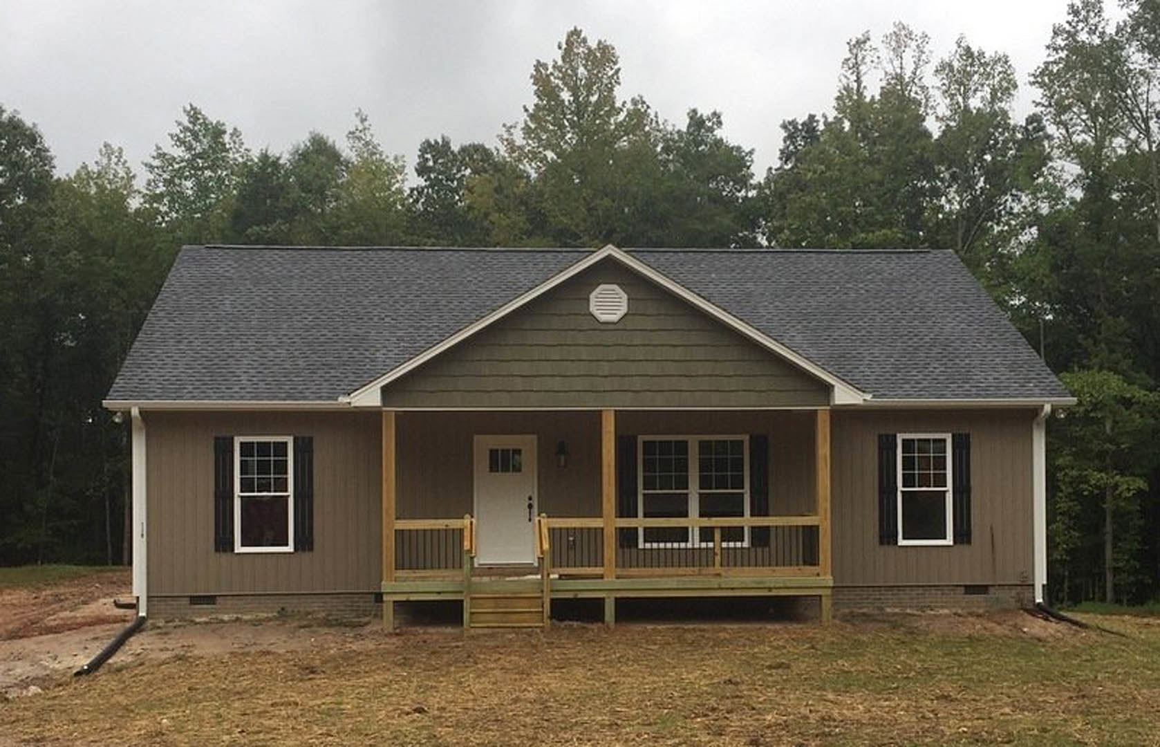 White cottage with horizontal siding, covered front porch, white door with window, railing, and roof vent; mature trees and blue sky in background.