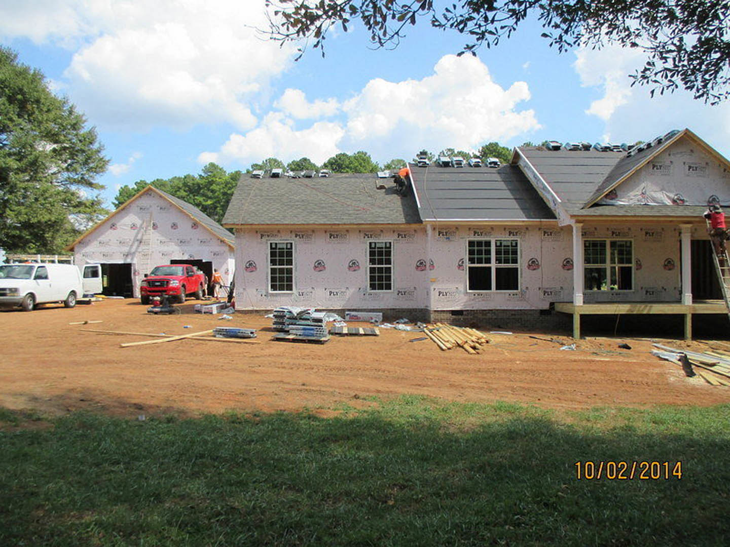 Partially built house with installed roof and grid windows, red truck and white van parked on street, leafy tree in foreground, cloudy sky overhead