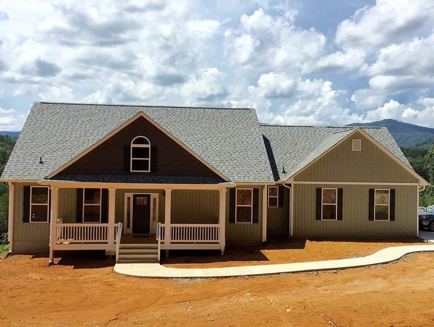 Two-story home under construction with white siding, black glass-paneled door, covered porch, white-framed windows, dirt road in foreground, blue sky with scattered clouds overhead