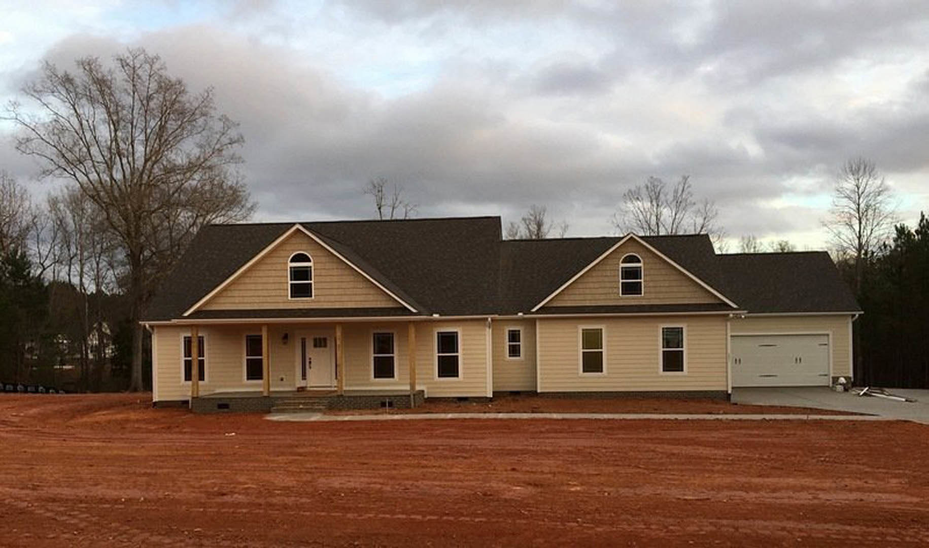 Two-story house with dark roof, white-framed square windows, surrounded by trees, situated next to a dirt road under a cloudy sky
