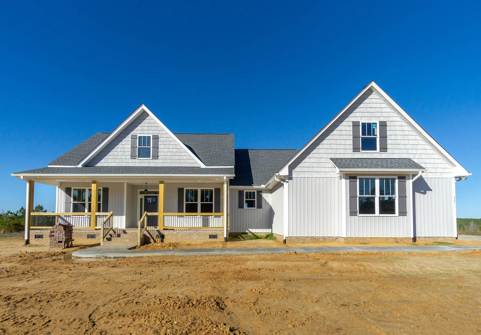 Two-story house under construction with white-framed windows, unfinished porch, exposed siding, and dirt yard bordered by a curb