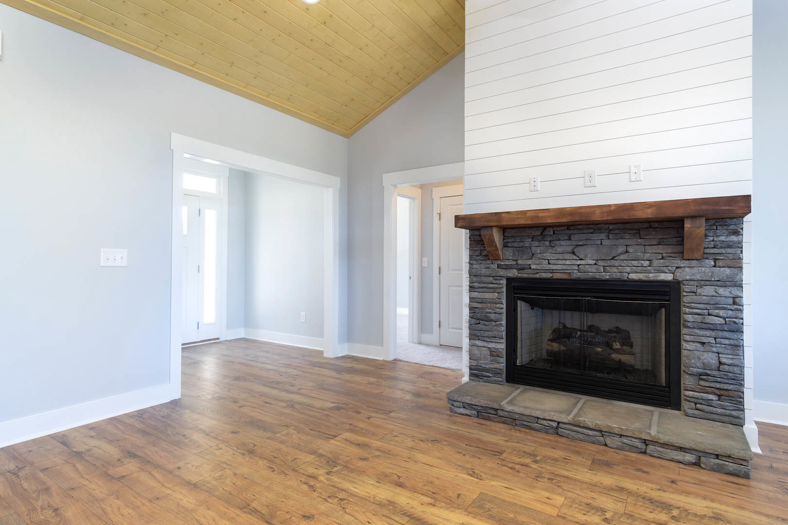 Stone fireplace with wood mantel, burning log, hardwood floors, white walls, three-switch outlet, and white door in bright living room