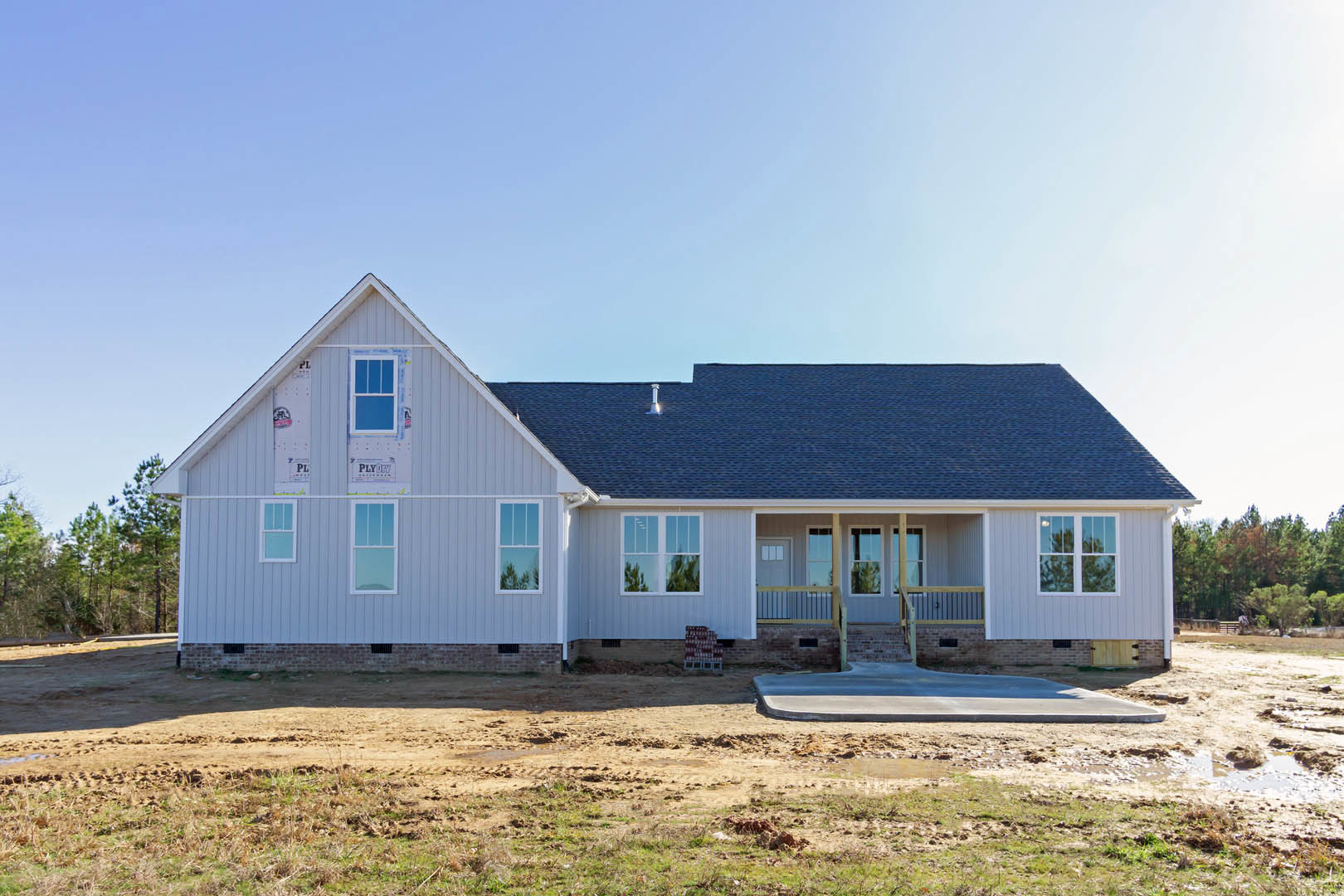 Two-story house under construction with exposed framing, dirt patch in front yard, stacks of building materials on pallets, covered porch, unfinished roof, and clear blue sky