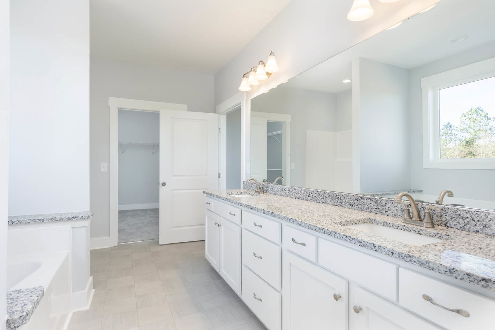 Bathroom with marble countertop double vanity, large wall mirror, silver faucets, white cabinetry and shelving, three-light fixture above, window with tree view.