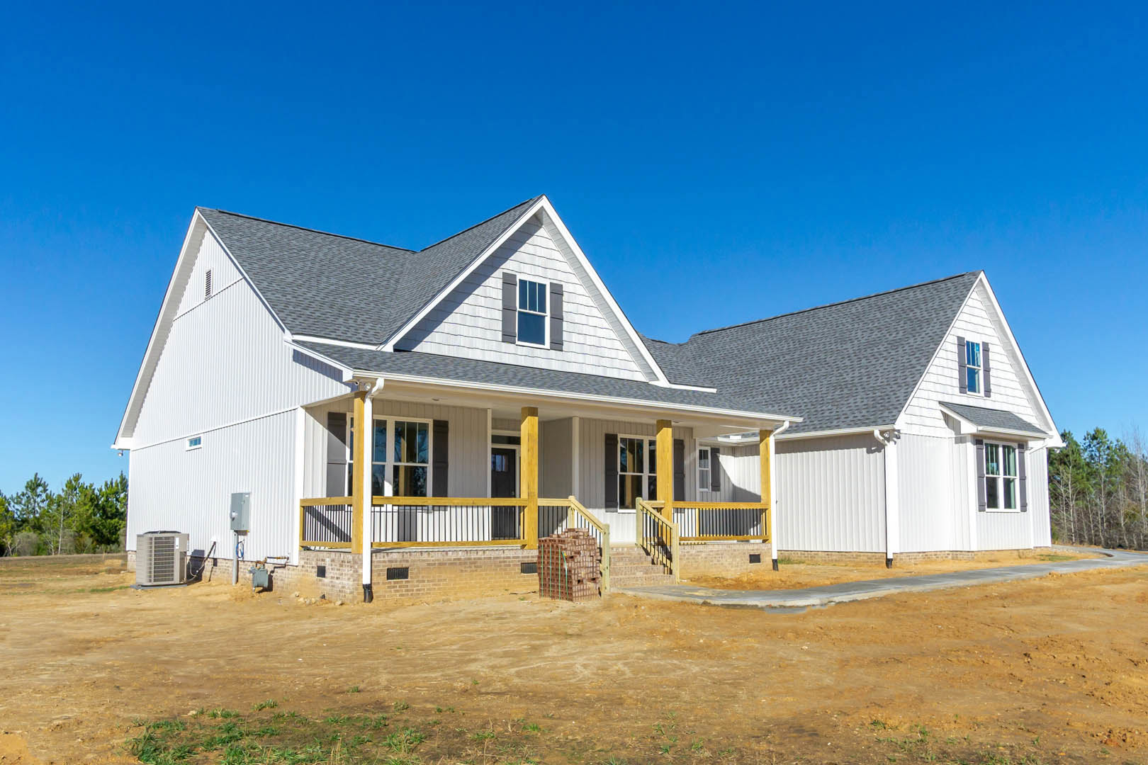 Two-story house with covered porch, white railing, multiple windows, dirt yard with stacked bricks on pallets, brick wall and stairs, blue sky overhead.