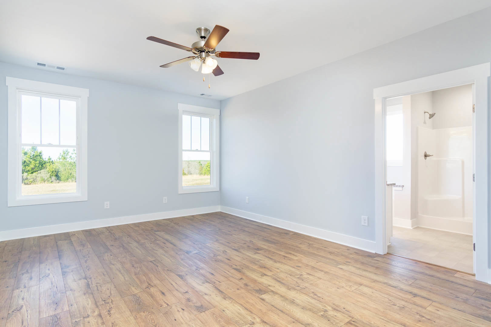 Ceiling fan with lights mounted on white plaster ceiling, wood flooring throughout room, large window with view of trees, partial view of bathroom shower in background