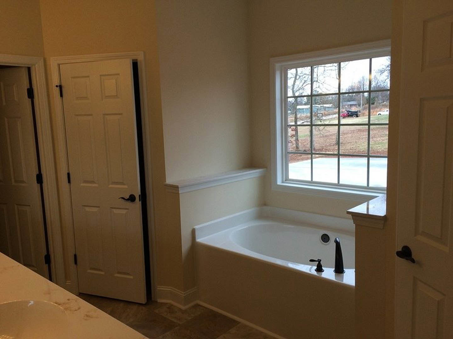 Bathroom with freestanding white bathtub, black faucet, large window overlooking yard and trees, white door with black handle, white countertop with footprints, black hat hanging