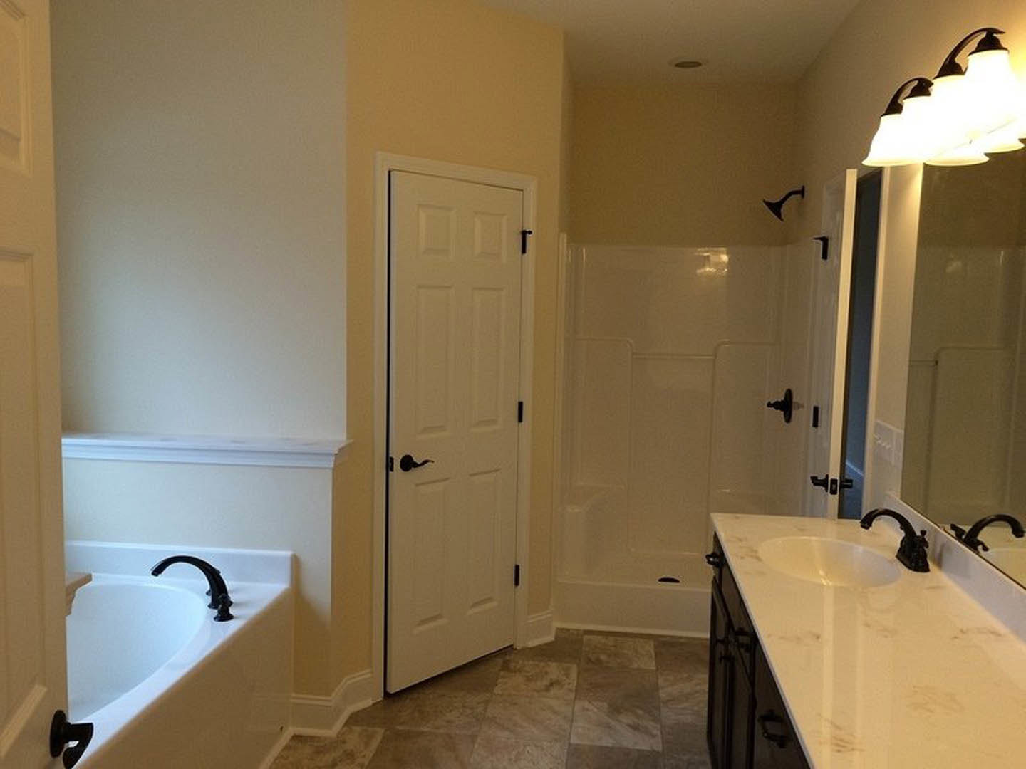 Bathroom with freestanding bathtub, white vanity with sink and chrome faucet, white door with black handle, wall-mounted row of lights, black towel hook, light-colored tile