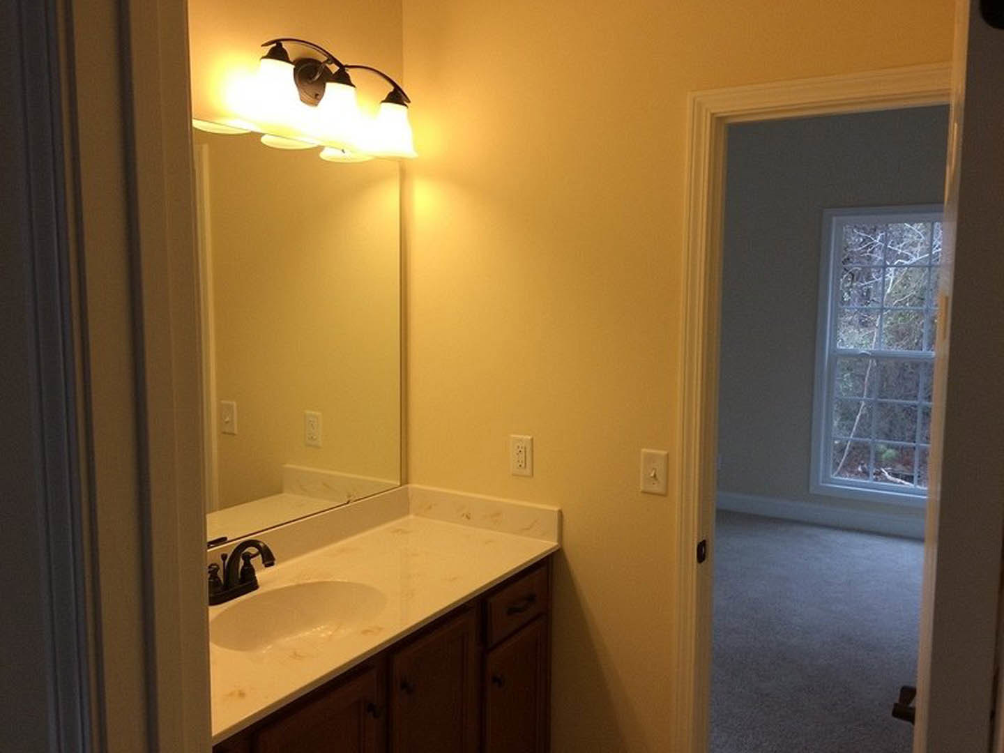 White quartz countertop with undermount sink, brushed nickel faucet, rectangular framed mirror, three-light fixture above, white tile backsplash, window with view of green trees