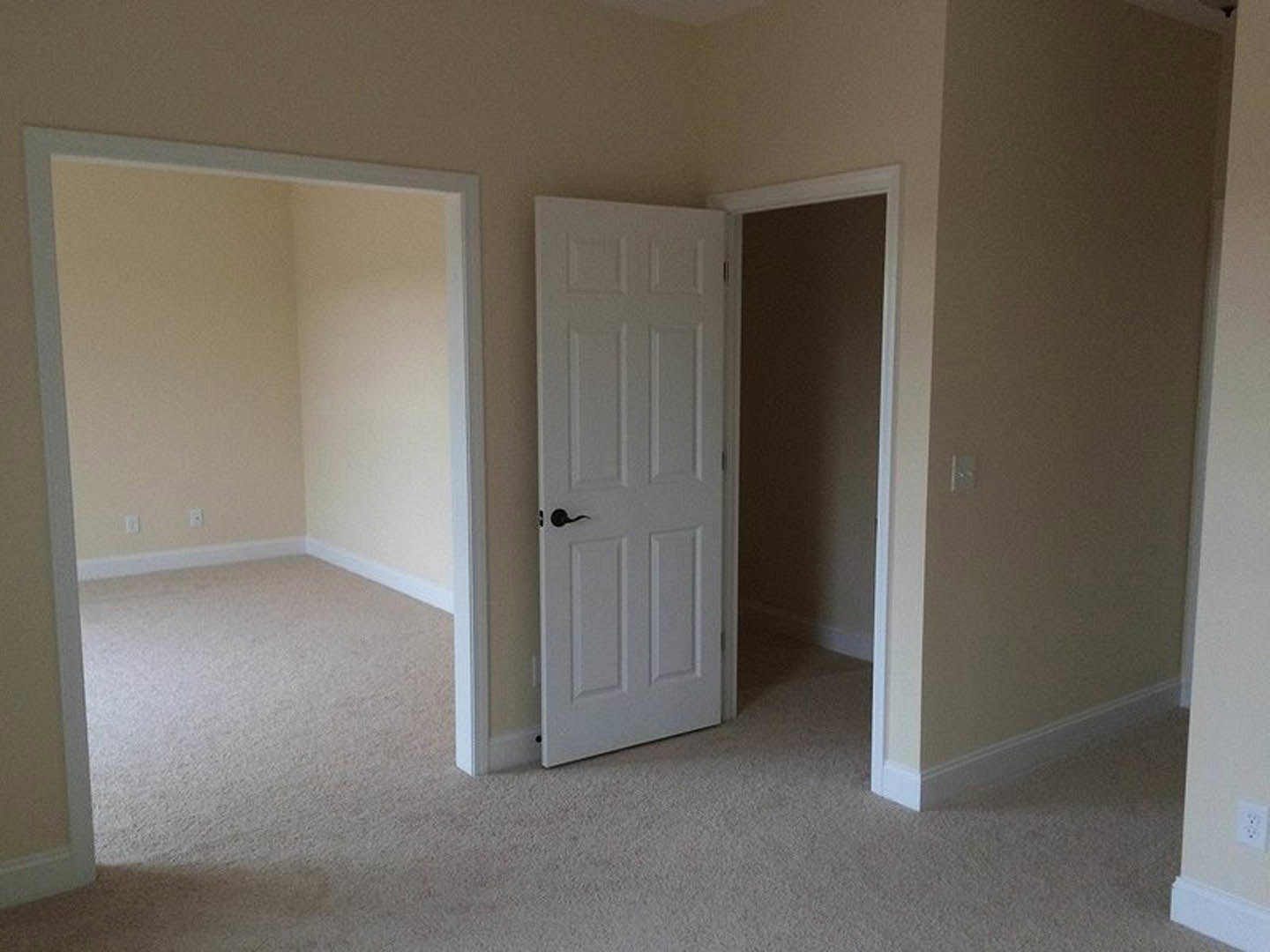 White paneled door with black handle set in a beige-walled room with light carpet, white door frame, and nearby light switch.