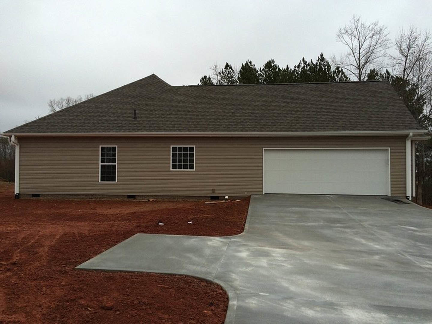 Concrete driveway bordered by dirt leads to a light-colored home with black trim, multiple square windows, and white-framed windows; trees and sky visible in background.