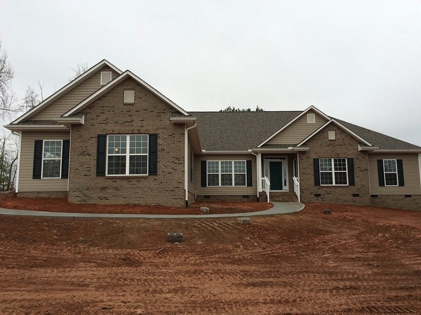 Two-story house under construction with gray siding, blue front door, multi-pane white-framed windows, dirt driveway, and scattered concrete blocks in front yard