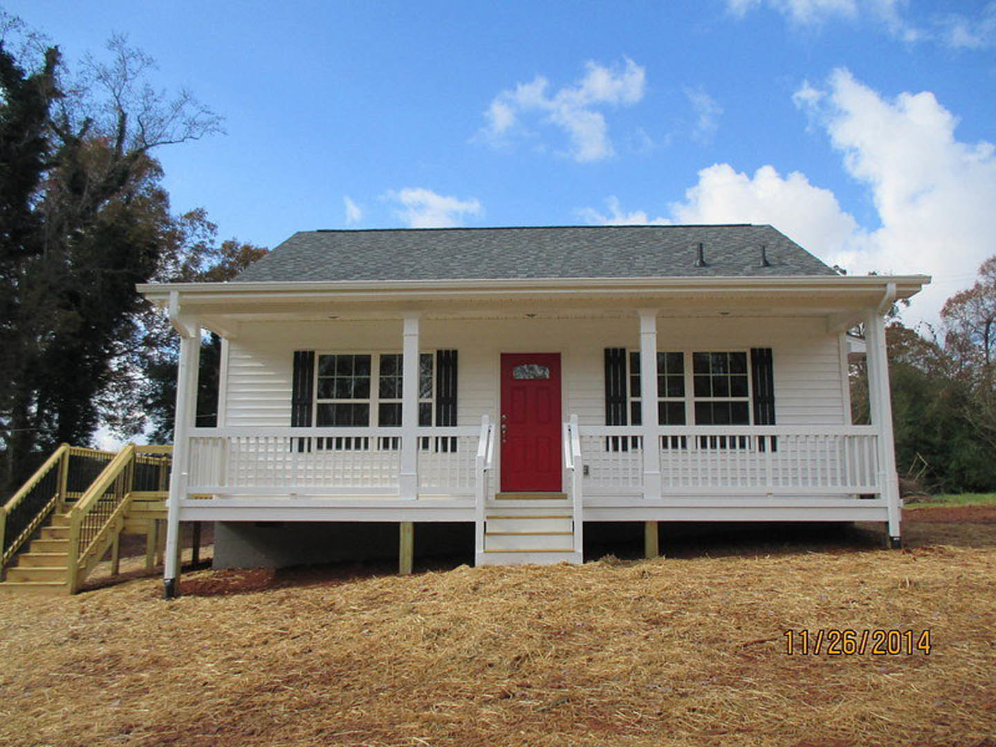 White siding exterior with a red front door featuring silver knobs, porch area, large window, tree nearby, and hay scattered on the ground