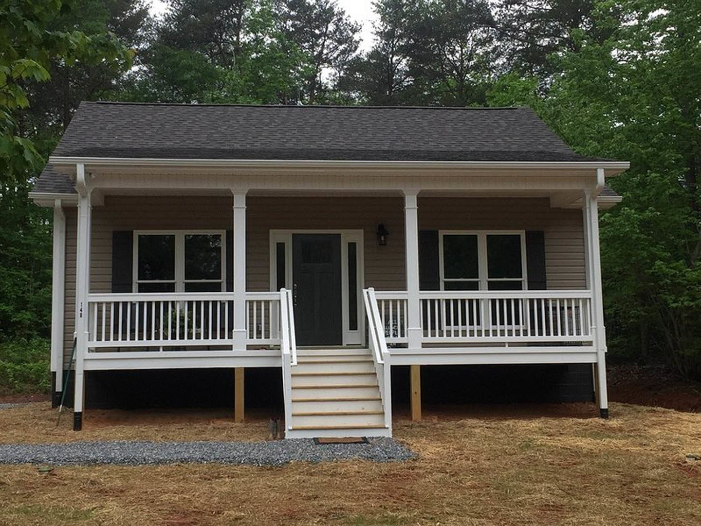 Two-story house with gray siding, black front door with white trim, white porch railing, covered porch, and stairs leading to entry, surrounded by trees and landscaping.