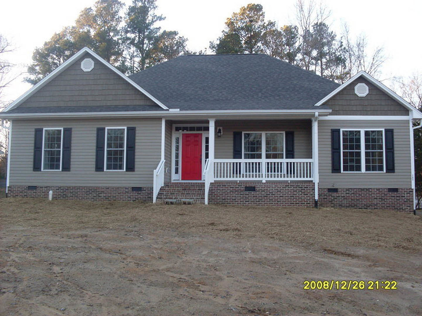 Red front door with white trim, grey siding, white vent, white porch railing on brick wall, window with white trim, tree and sky in background