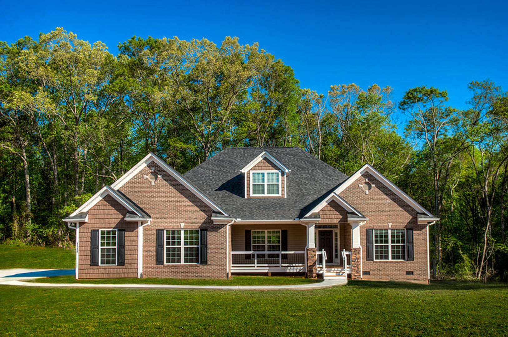 Two-story home with white siding, black front door, covered porch featuring white railing and chair, large windows, gabled roof with dormer window, surrounded by grass and mature