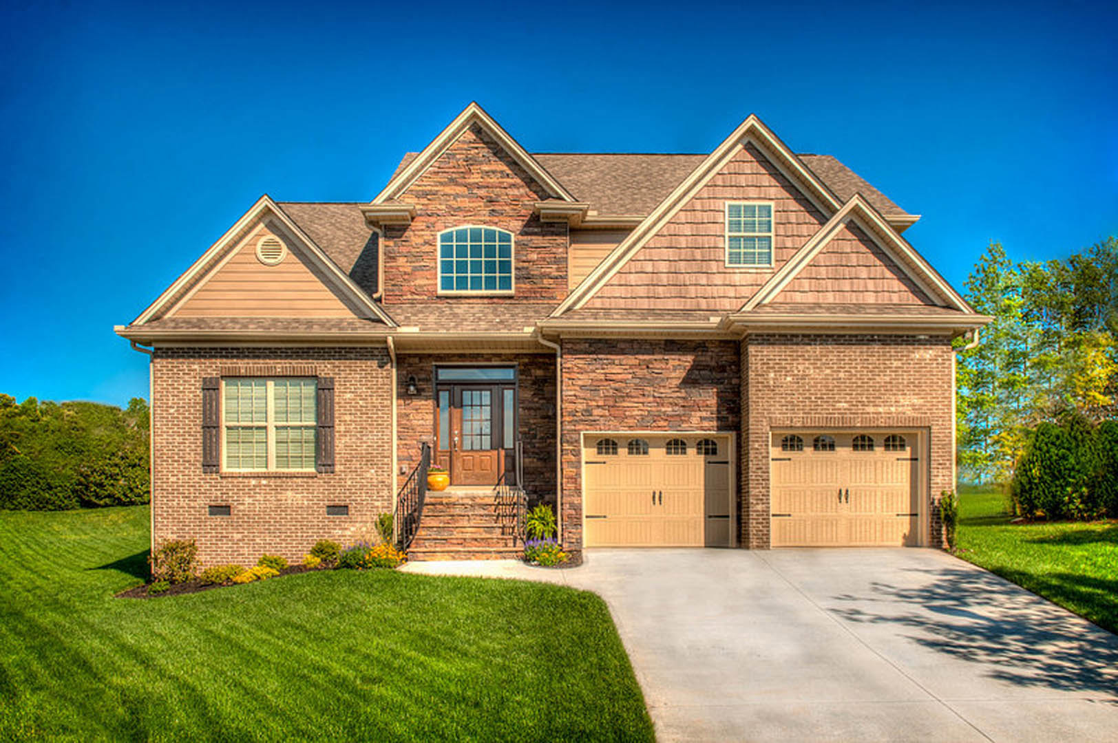 Two-story house with white siding, large windows, attached garage, concrete driveway, green lawn, and tree shadows cast across driveway and sidewalk