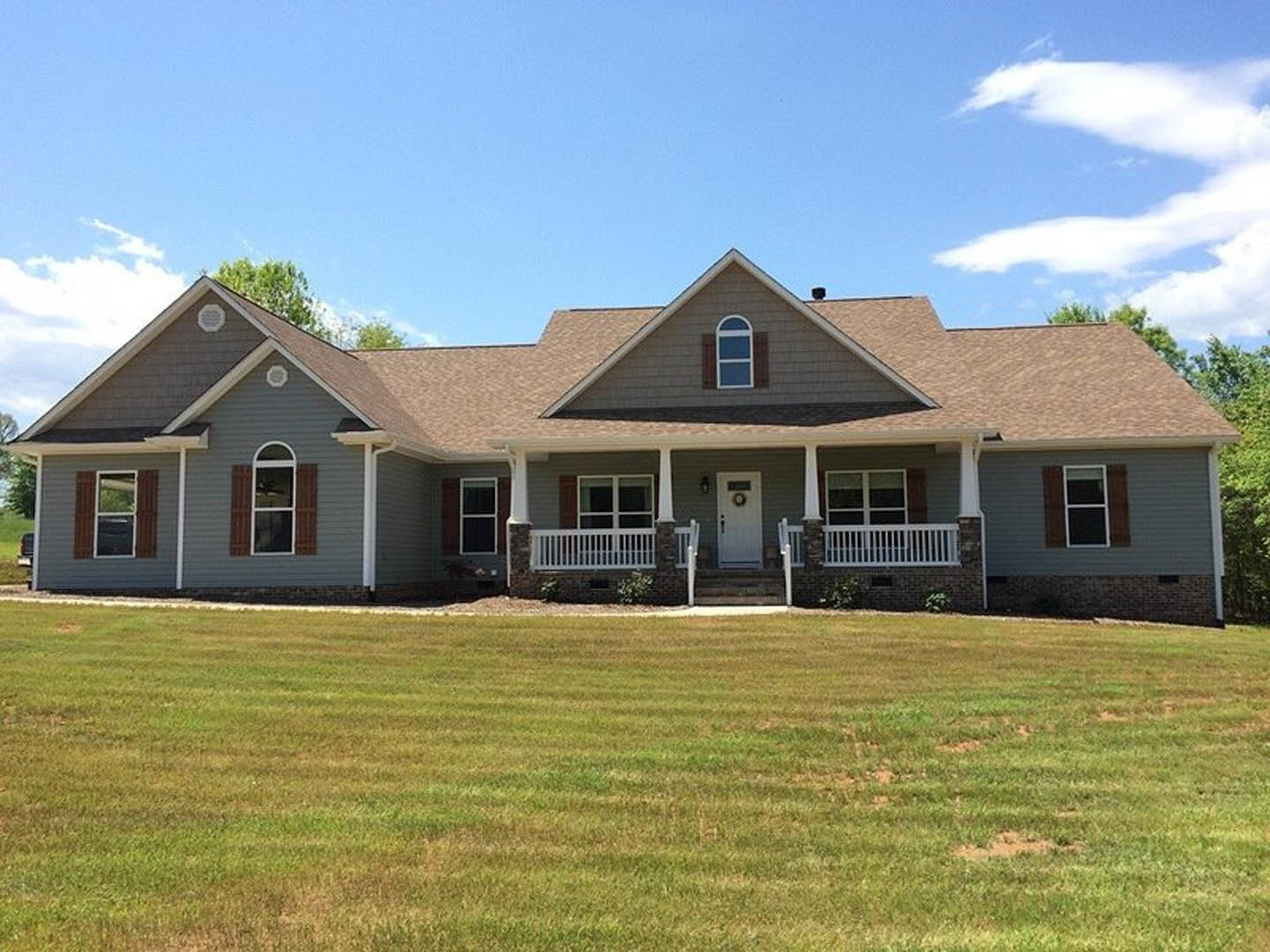Two-story home with white porch railing, white-framed windows, and a wreath-adorned front door, set behind a large green lawn under a partly cloudy blue sky