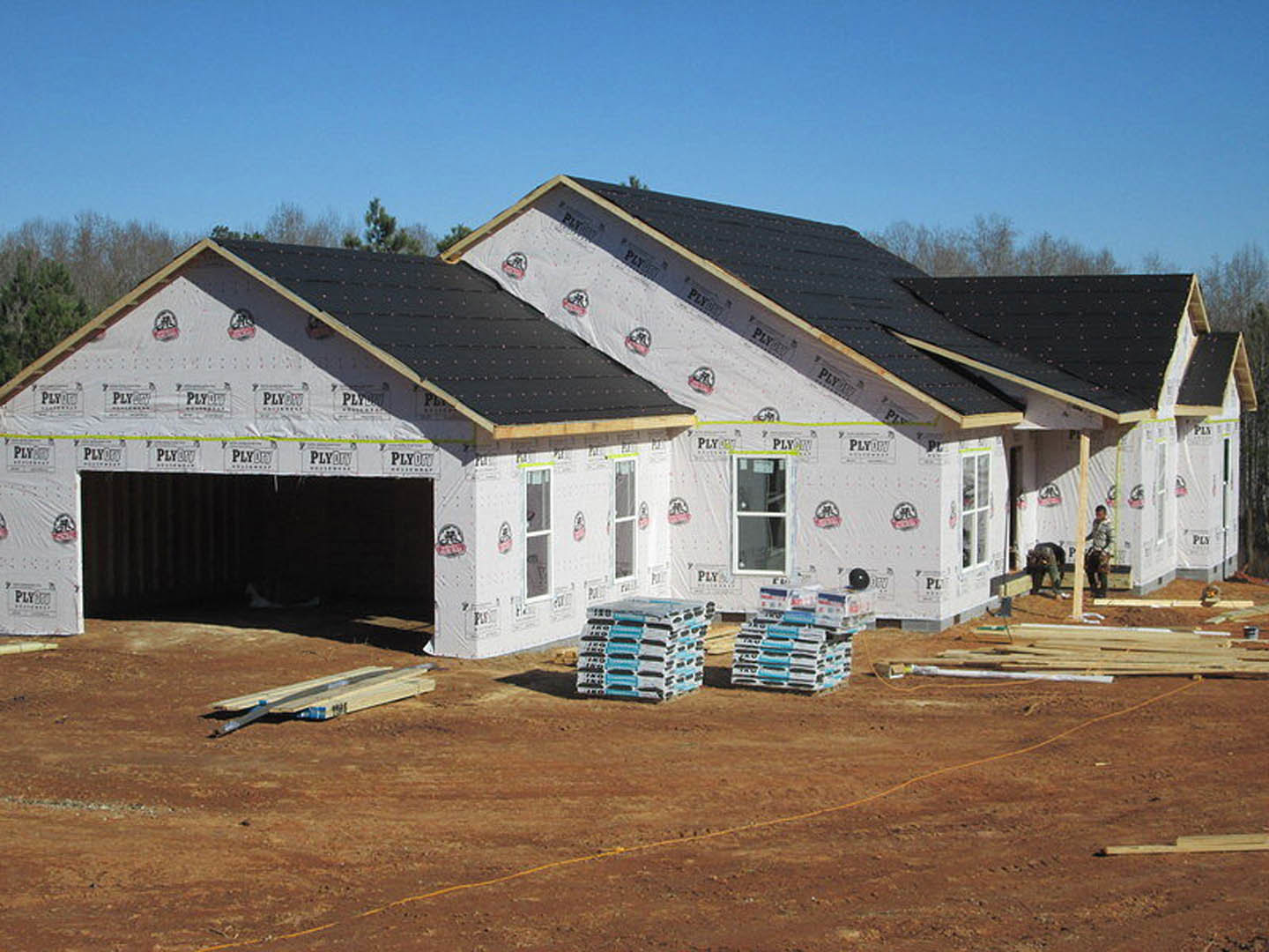 Partially built house with black roof, exposed framing, stacks of cement bags, wooden planks, and white and blue pallets on dirt ground