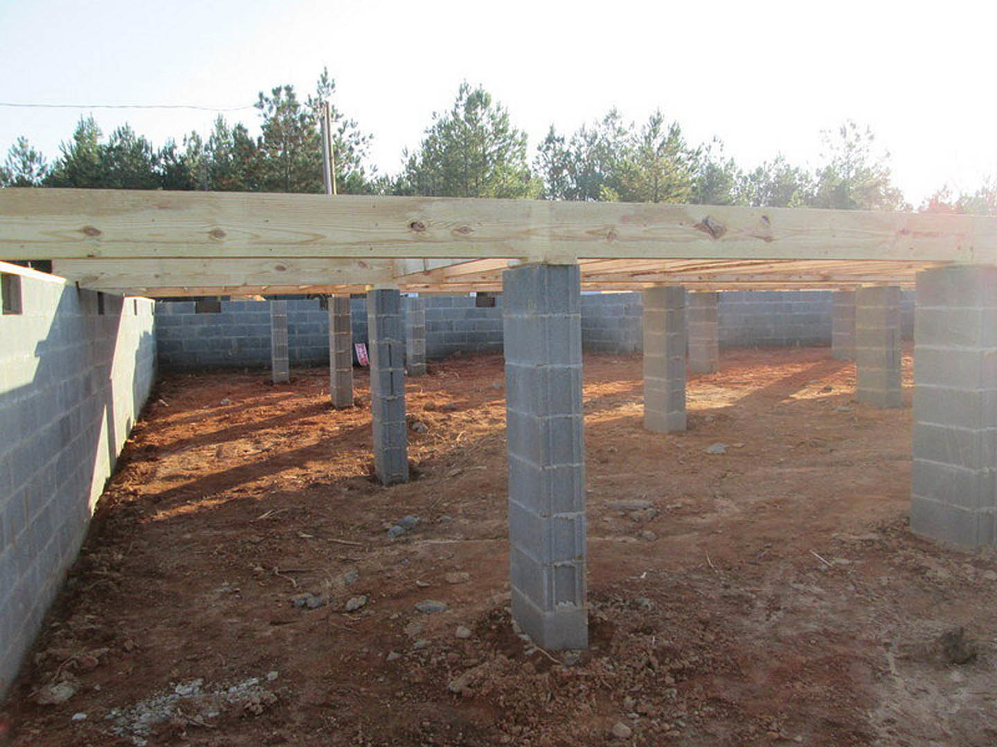 Concrete foundation with square pillars, wooden beams framing the structure, white brick wall partially built, construction materials scattered on bare ground, exterior view with