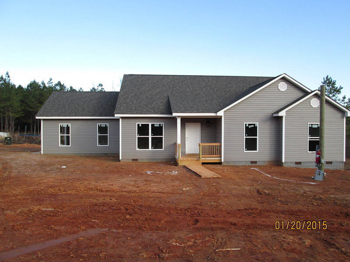 Partially built house with exposed wooden framing, white front door, covered porch, and red dirt yard under clear sky