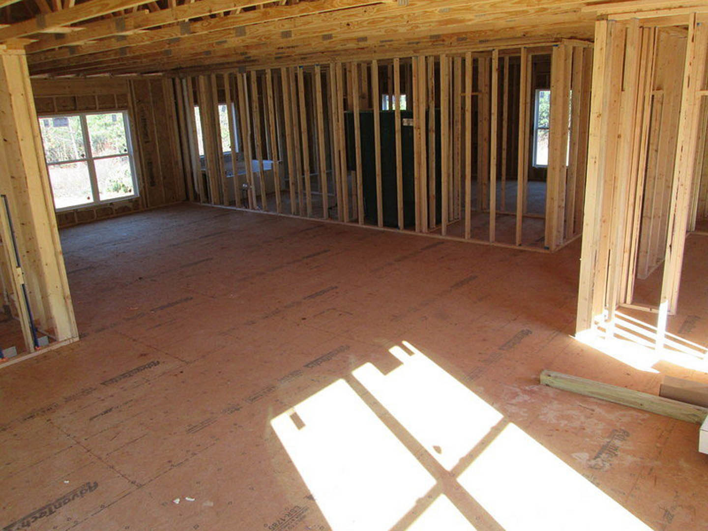 Wood-framed room with exposed beams, hardwood floor, white-framed window, and wooden ceiling columns; sunlight casts shadows across the floor.