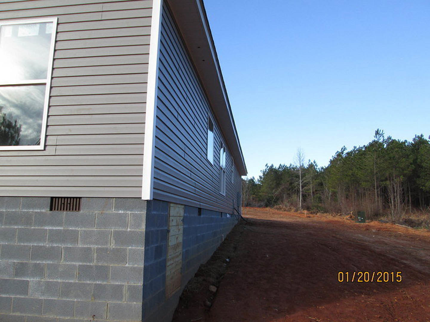 Partially built brick house with white-framed windows, surrounded by dirt and trees, construction materials visible near exterior siding