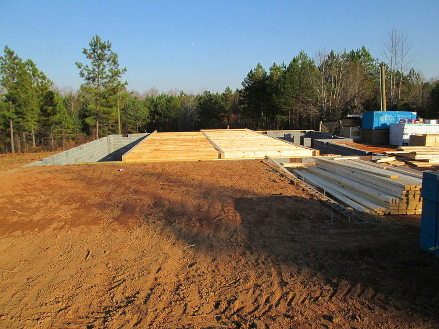 Dirt construction site with tire tracks, stacked lumber, partially built wooden stairs, leafy tree, and blue sign with white text