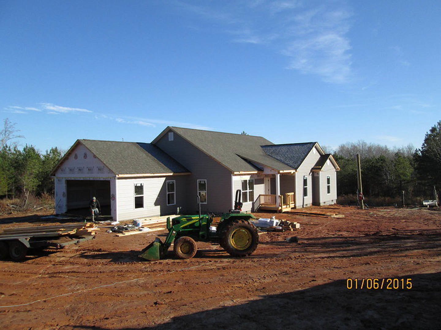 Green tractor with front bucket parked on dirt driveway in front of partially constructed house, blue sky overhead, trees in background.
