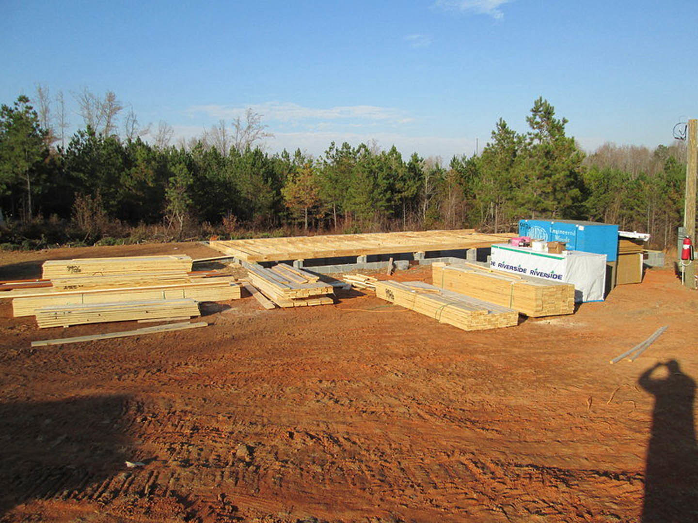 Wood planks stacked on dirt at a residential construction site, blue shipping container in background, scattered tools and materials, trees and cloudy sky visible