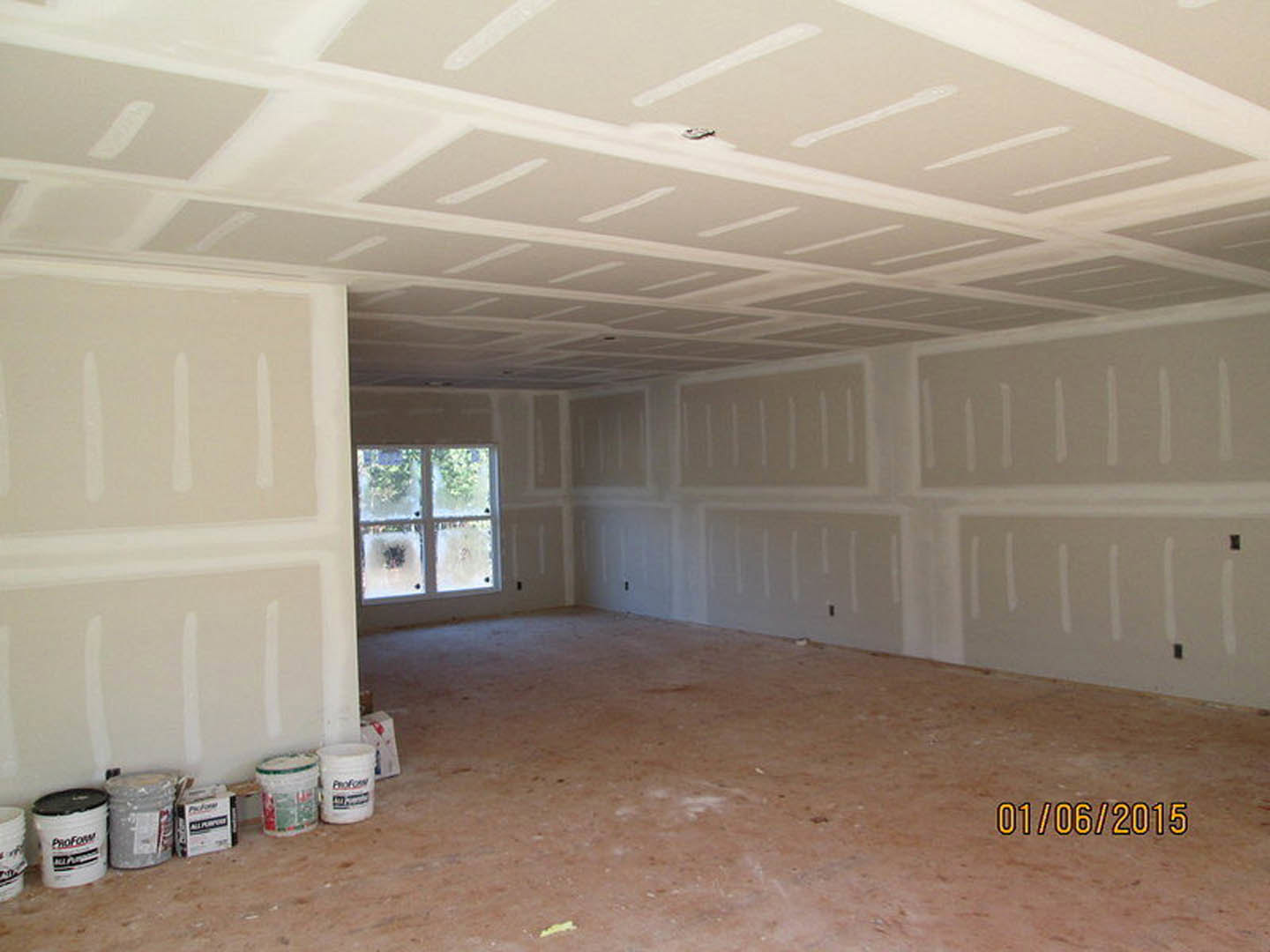 Unfinished room with plaster walls, dirty floor, paint cans, white bucket with black lid, frosted glass window, exposed ceiling beams