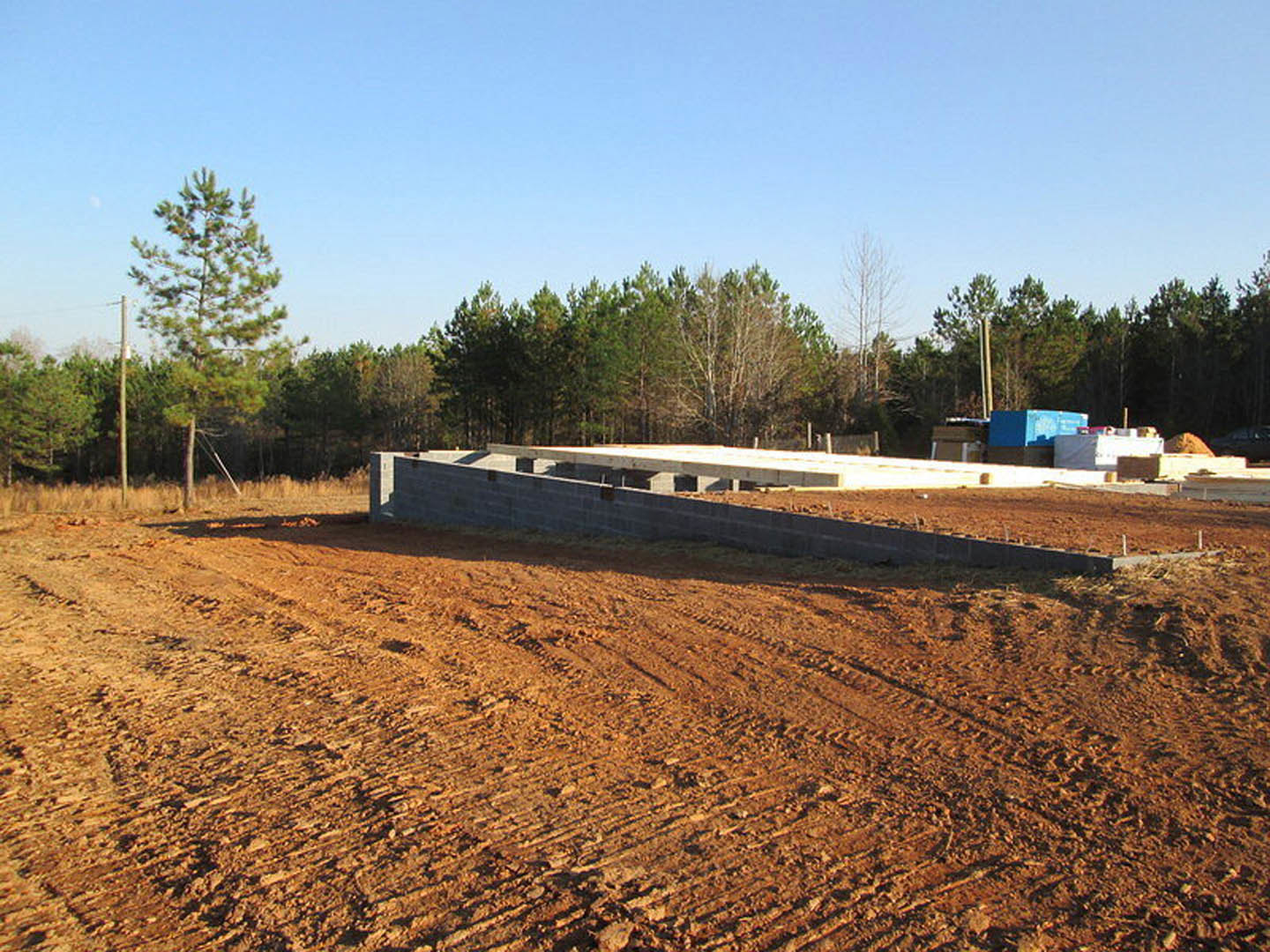 Concrete foundation and block wall on dirt construction site, surrounded by trees with green leaves under blue sky