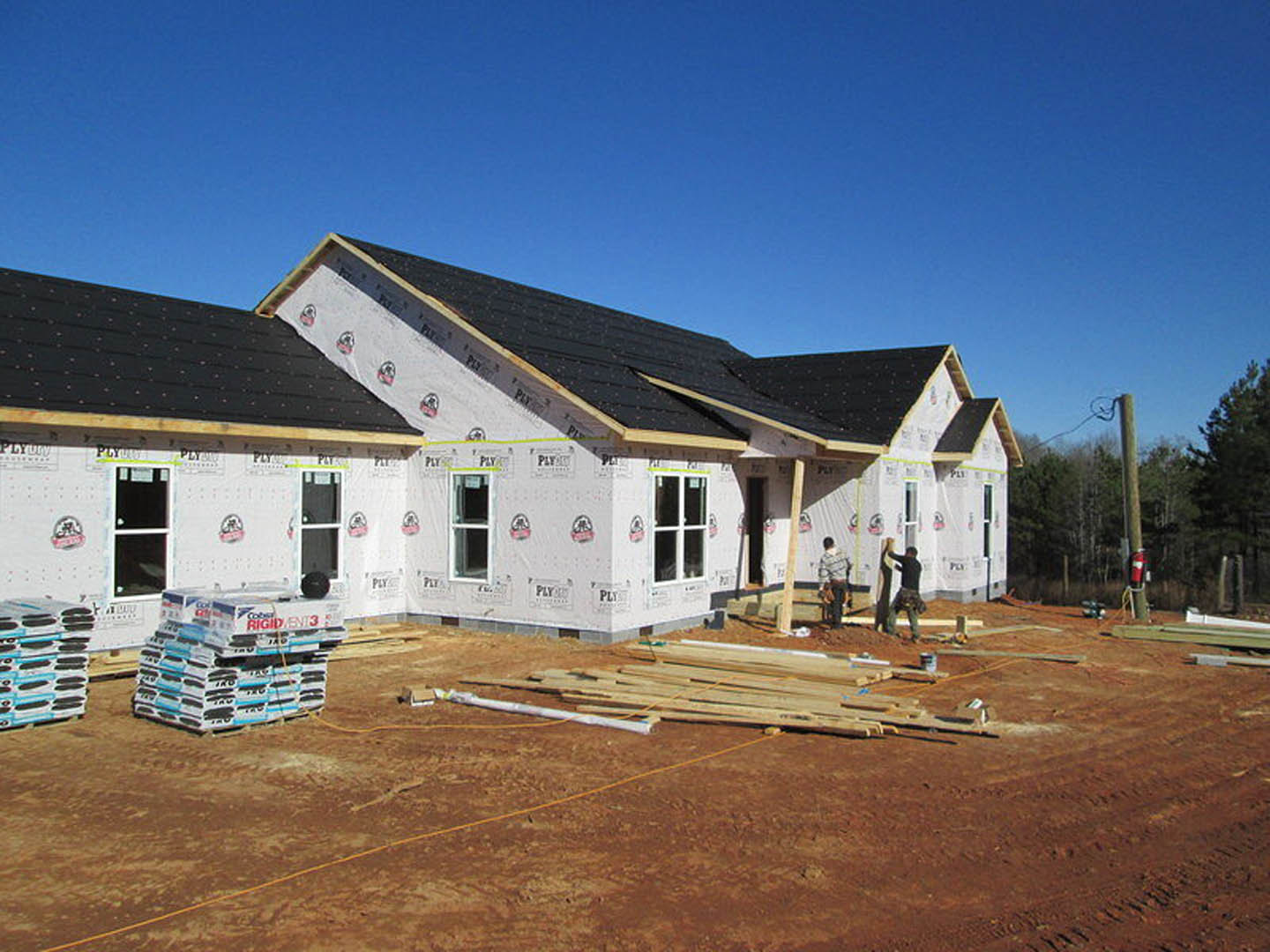 Partially built home with exposed insulation, construction materials including stacked boxes and wood on the ground, several workers present, unfinished exterior walls, visible