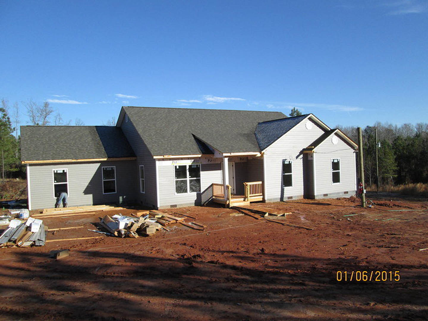 Partially built house with exposed framing, porch area, wooden bench and railing, pile of lumber on ground, several people working, square-paned window visible, surrounded by trees
