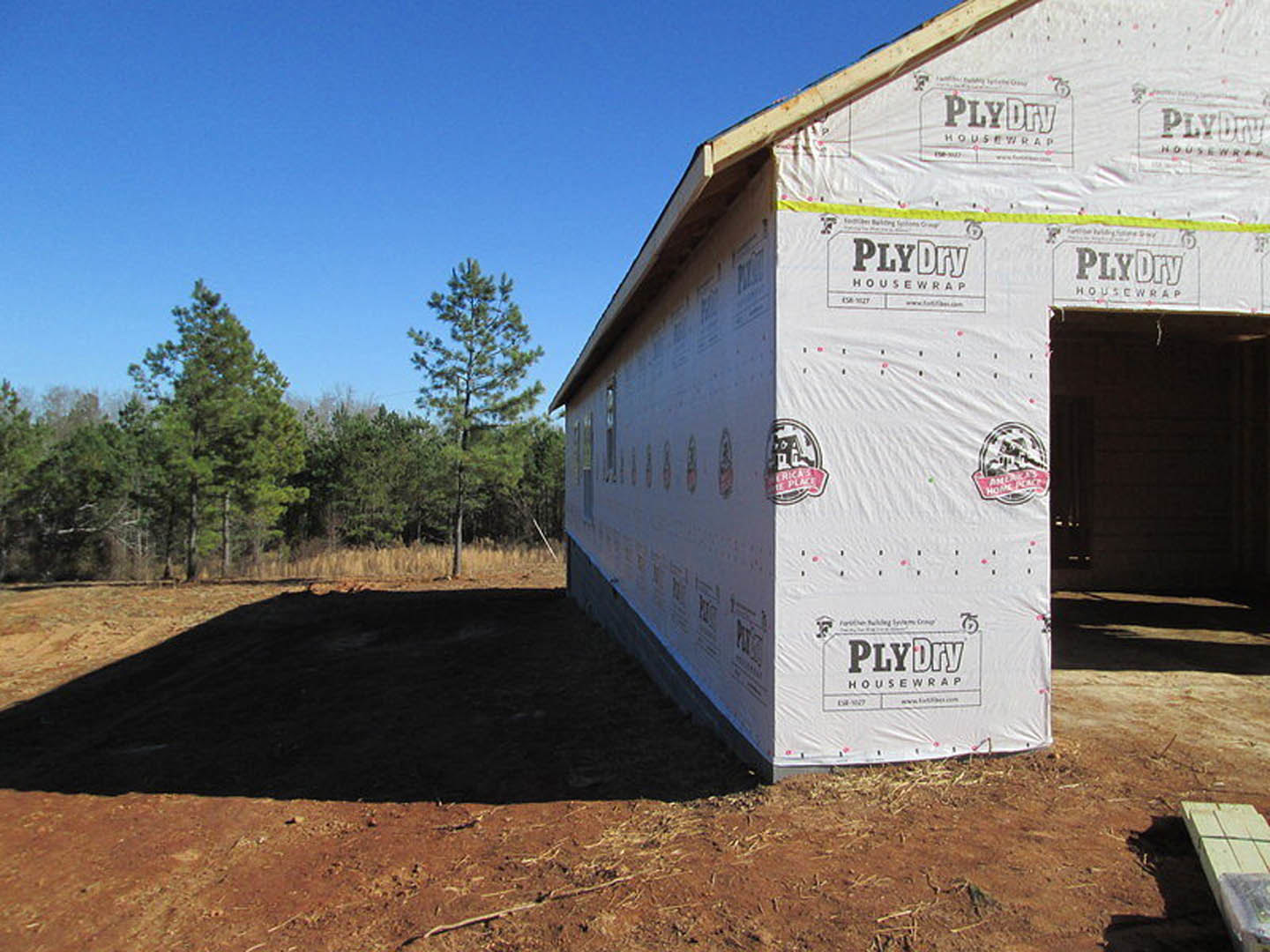 Partially built house wrapped in white weatherproof sheeting, surrounded by green trees and grassy yard, construction materials scattered near foundation, temporary sign posted in