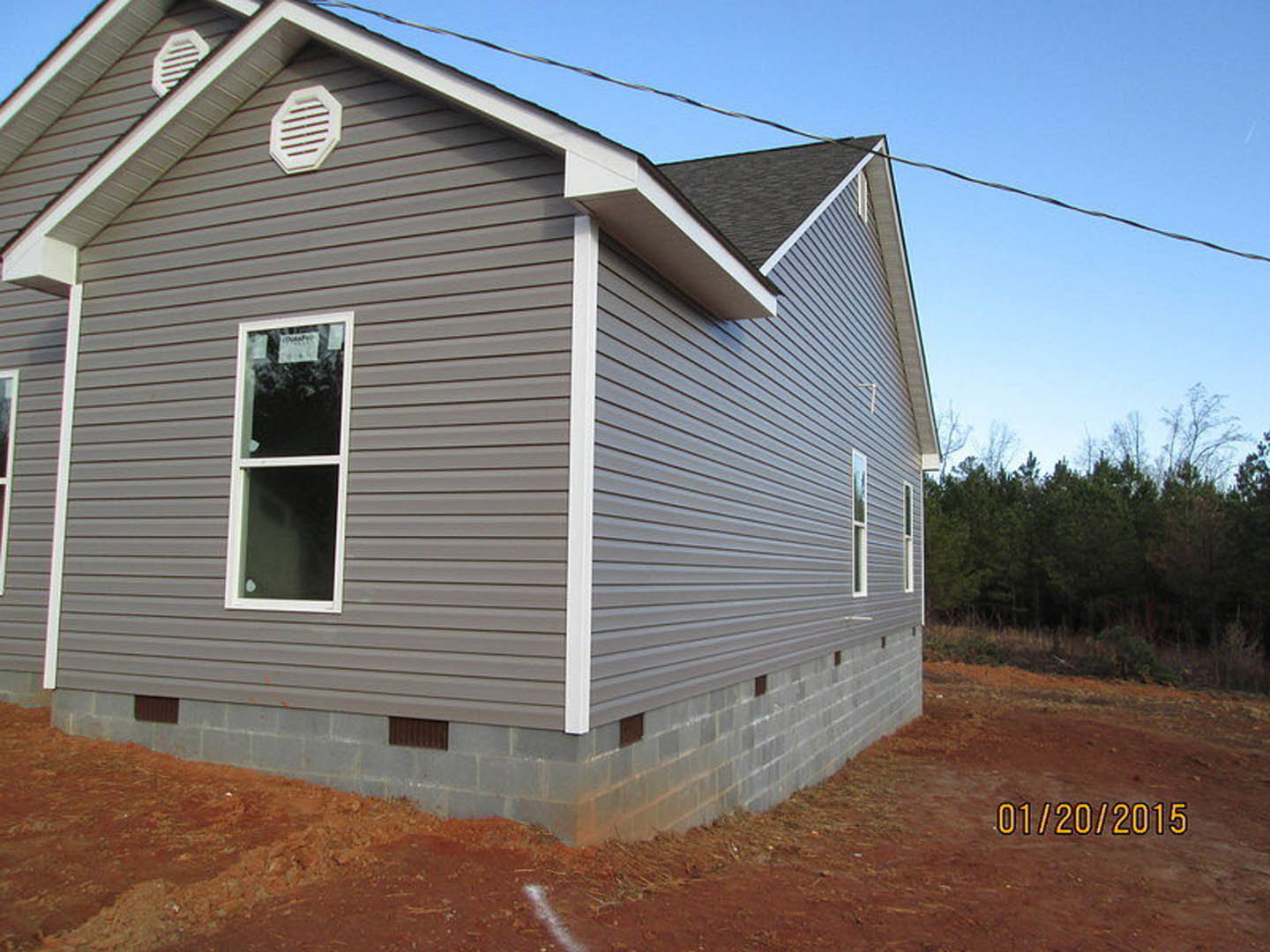 Framed house under construction with exposed wood, broken window glass, white wall vent, overhead power line, and blue sky; trees visible in background.